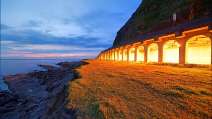 Scenery of a coastal highway by rocky beach before sunrise with lights from the rock shed tunnel illuminating the coast ~ Beautiful view of a beach north to Taipei, Taiwan in morning twilight