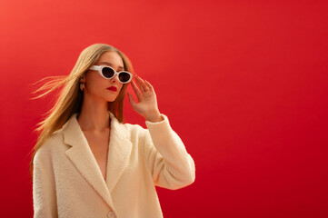 Fashionable confident woman wearing trendy sunglasses, white boucle coat, posing on red background....