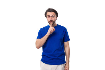 young brutal caucasian brunette man with a beard in a blue t-shirt keeps the secret of business development on a white background with copy space