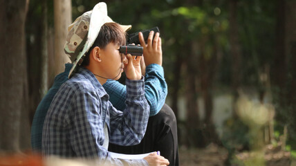 Young Asian boys are using a binocular to lookout for birds and animals in a local park, soft and...