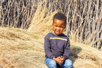 african village , child sitting on thatched grass in the yard by the fence