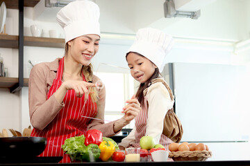 Happy Asian mother and daughter girl wear apron chef hat at kitchen, mom touch transparent device tablet for searching tasty meal recipes on internet, cute kid chef and family cooking food at home.