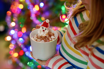 Little child girl holding cup with hot chocolate with marshmallows as Santa Claus. Kid by near Christmas tree decorated with lights.