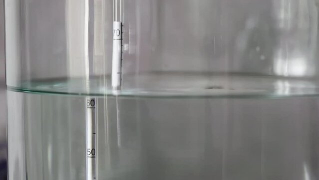alcohol distilling process closeup - liquid distilled transparent liquid flowing in large glass jar with floating alcoholometer in it.