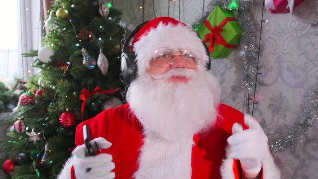 Fiery dancing Santa Claus with beard and glasses in modern phone and headphones dances and waves arms against  background of Christmas decorations, spreading festive mood and giving gifts. Close up.