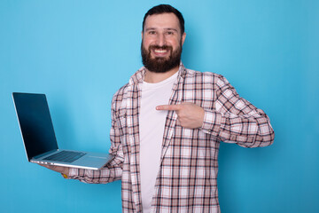 Smiling bearded man in casual clothes posing isolated on blue background, studio portrait. People lifestyle concept. Mock up copy space. Pointing index finger on laptop pc computer