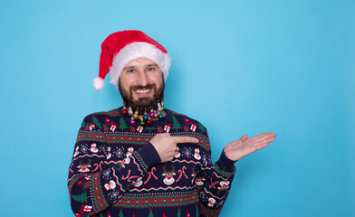 Handsome caucasian bearded man in red Christmas cap pointing to free space and looking at camera isolated over blue background. Christmas sale.