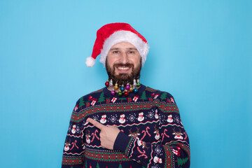 Obraz premium Handsome caucasian bearded man in red Christmas cap pointing to free space and looking at camera isolated over blue background. Christmas sale.