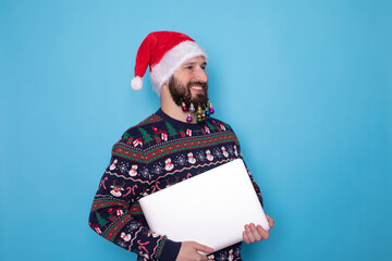 Portrait of handsome young bearded male hold laptop working remotely wear x-mas print vest hat garment isolated on blue background.