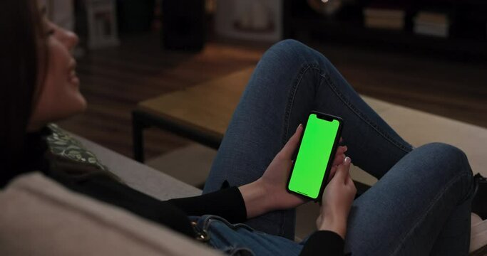 Female sitting on couch smiling laughing holding cell phone with green mock up screen in hands watching video movie film observing photos using mobile phone internet social networks browsing.