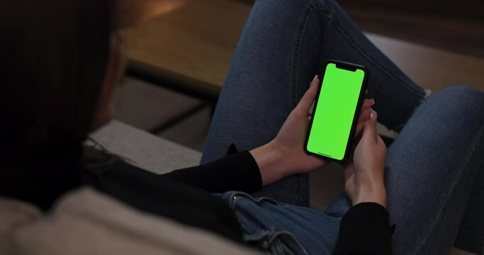 Yong btunette woman in the living room sitting on couch sofa holding smartphone cell phone with green mock-up screen. Girl surfing Internet observing images video.