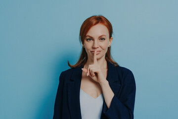 Redhead woman with finger on lips signaling silence, wearing a navy blazer, against a blue...