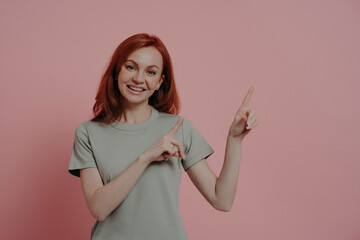 Smiling redhead woman pointing upwards with her finger, dressed in a casual tee on pink