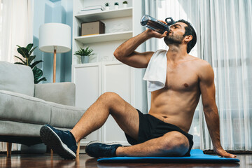 Athletic and sporty man drinking water on fitness mat after finishing home body workout exercise...
