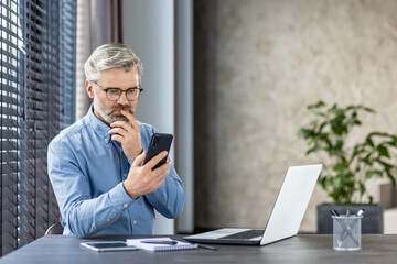 Serious thinking mature gray haired man at workplace inside office, businessman holding phone, thoughtfully reading message browsing email and social media.