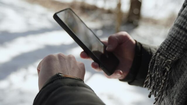 European man in warm clothes synchronizes smart watch with smartphone and taps on screen in winter, closeup. View statistics from fitness bracelet, configure it during health walk on frosty sunny day.