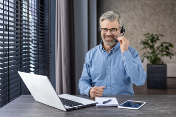 Portrait of mature successful gray haired man inside office at workplace inside office, businessman with headset phone and laptop for video call smiling and looking at camera.