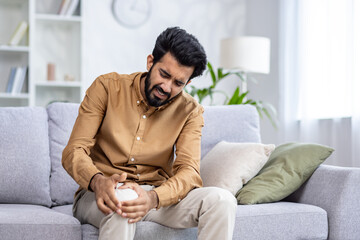 Indian young man sitting on the sofa at home and holding his knee, suffering and grimacing in pain.