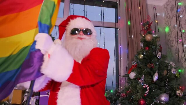 Modern Santa Claus wearing black glasses fiercely supports the LGBT movement waving and dancing rainbow flag at Christmas party decorated with festive tree with glowing garlands, wreaths and tinsel.