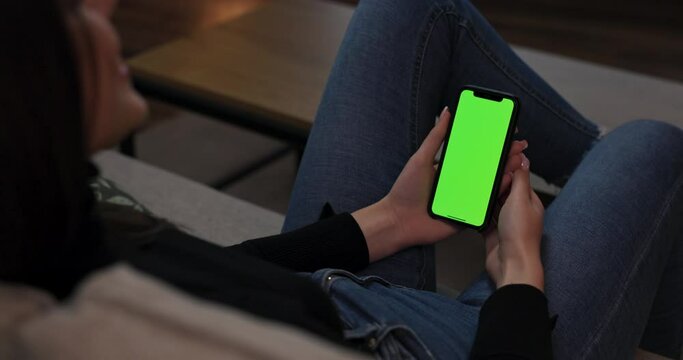 Mock- up image young brunette woman in casual outfit sitting on couch holding cell phone with green mock-up screen. Point of view shot.