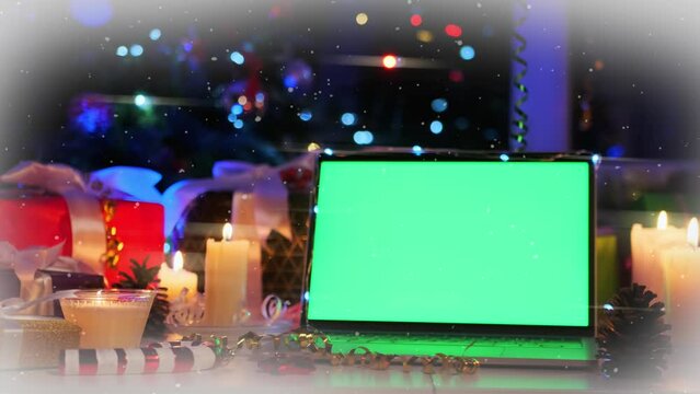 Portable laptop with green screen, xmas decoration of glowing candles, wrapped gifts and flickering multicolor bokeh lights on snowy background. Holiday offer place.