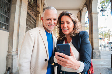 Fototapeta premium Mature tourists couple using a smartphone and watching funny pictures on their weekend trip and enjoying together. Middle aged woman and her husband searching hotel on a cell phone standing outside