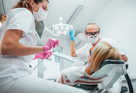 Dental clinic patient appointment. Dentist man in magnifying glasses pointing light. Young female assistant ready for teeth surgery with medical tools. Health care and medicare industry concept image.