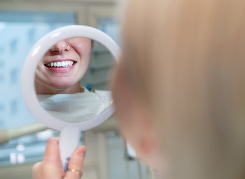 Young smiling woman sitting in stomatology clinic chair and looked at mirror evaluating her teeth reflection after Tooth whitening procedure. Health care and medcare industry concept image