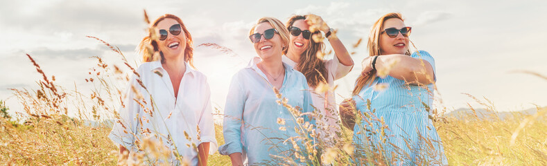 Portrait of four cheerful smiling and laughing women during outdoor walking by a high green grass hill. They looking at the camera. Woman's friendship, relations, and happiness concept image. - obrazy, fototapety, plakaty