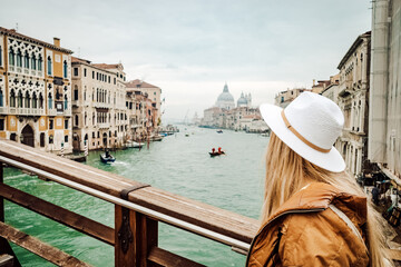 Fototapeta na wymiar A stylish woman gazes upon the bustling cityscape and tranquil river, adorned in a fashionable hat and clothing, as a ship and gondola glide by on the water