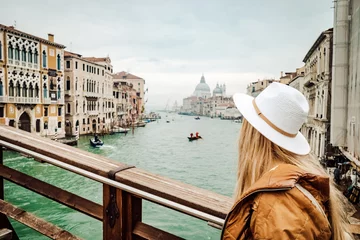 Tuinposter A stylish woman gazes upon the bustling cityscape and tranquil river, adorned in a fashionable hat and clothing, as a ship and gondola glide by on the water © Armen