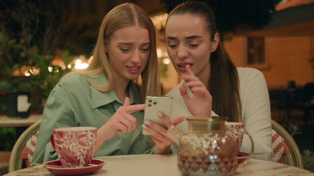 Two European Caucasian women drinking tea elegance cafe using phone smartphone online chat arrogant pathos skeptical condemn city outside best friends disapproval judgment reaction gossip expression