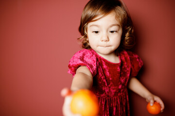 Adorable caucasian brunette toddler little girl in red velour  holding mandarin oranges in hands at...