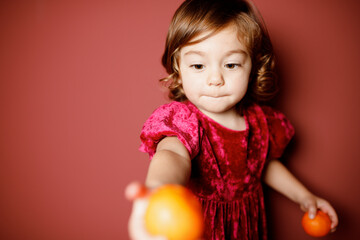 Adorable caucasian brunette toddler little girl in red velour  holding mandarin oranges in hands at the red background. Christmas vibe 