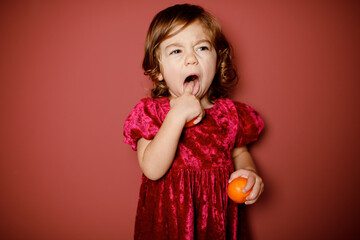 Adorable caucasian brunette toddler little girl in red velour with dissatisfied grimace as for...