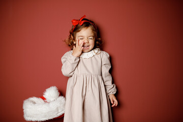 Adorable caucasian brunette toddler little girl in beige dress and red ribbon smiling widely with the finger in noise at the red background. Christmas vibe 