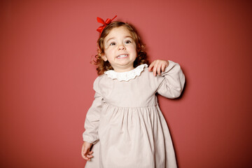 Adorable caucasian brunette toddler little girl in beige dress and red ribbon smiling widely at the red background. Christmas vibe 