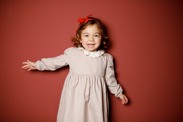 Adorable caucasian brunette toddler little girl in beige dress and red ribbon smiling widely at the...