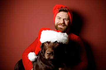 Portrait of Santa Claus bearded in red hat with chocolate Labrador retriever wearing red Santa hat...