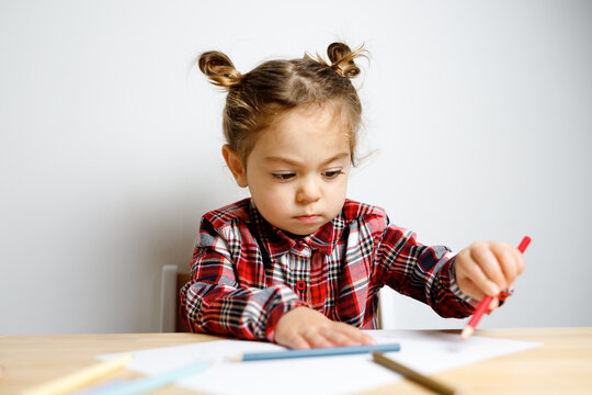 Portrait of cute toddler girl with two bunches in checkered dress sitting on the baby wooden table and chair and drawing with colorful pencils, Montessori concept fine motricity 