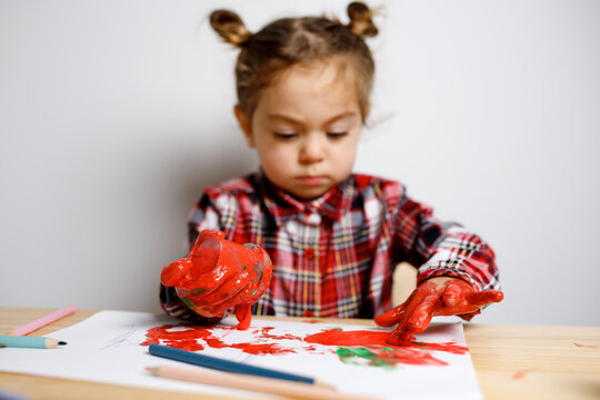 Portrait of cute toddler girl with two bunches in checkered dress sitting on the baby wooden table and chair and drawing with red finger paints with messy hands, Montessori concept fine motricity 