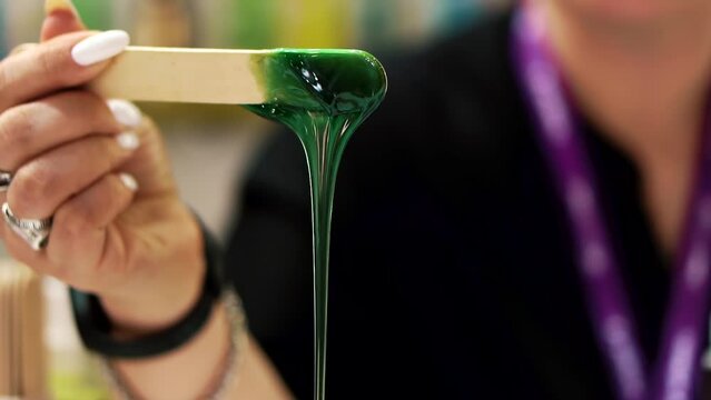 A woman's hand with rings on her fingers picks up a wooden stick from container. Viscous green sugaring paste drips from the end of the wooden stick. Concept of benefits of depilation for body beauty