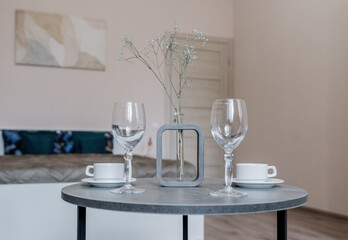 a round grey table. there are glasses on the table, white mugs and a vase with flowers. There is a bed in the background