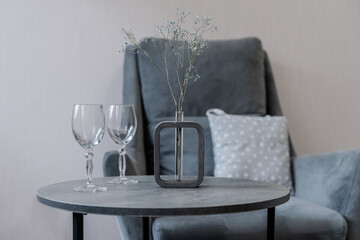 a round grey table. there are transparent glasses and a vase of flowers on the table. There is an armchair in the background.