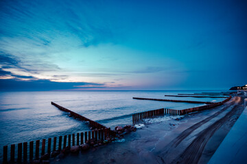 As the sky blazes with a fiery sunset, a rugged wooden fence guards the horizon of the vast ocean, with a solitary pier stretching into the wild seascape
