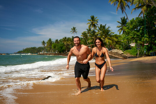Happy couple runs on tropical beach, man and woman jogging near sea, fit duo enjoys summer. Fit male, slender female in swimwear keep active, coastal lifestyle. Athletic pair smile, ocean fun.