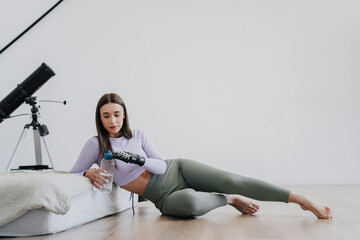 Beautiful caucasian girl in sportswear laying on floor at home opens bottle of water by hi tech...