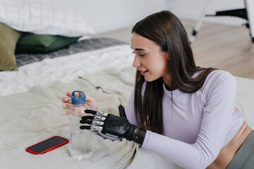 Young woman laying on bed  at home, with  bionic hand prosthesis after workout holds bottle with...