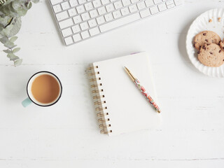 Break and snack in office work. Flat composition with copy space on white desk table with coffee cup and plate of cookies next to notebook and pen next to computer keyboard.