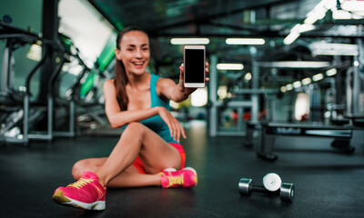 Fototapeta na wymiar Apps on smartphones for the gym. Close up photo of beautiful slim woman in sportswear sitting on the floor showing the phone on camera. Focused on phone